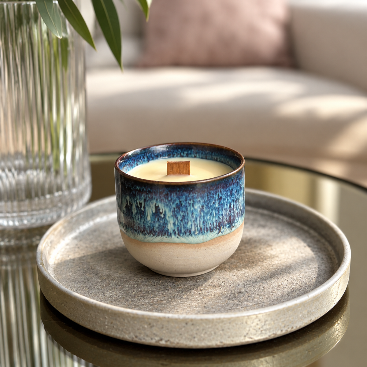 A ceramic candle in a small speckled gray tray on a glass table, with a blue and cream glazed finish and a wooden wick. A ribbed glass vase with green leaves and a blurred sofa are in the background.