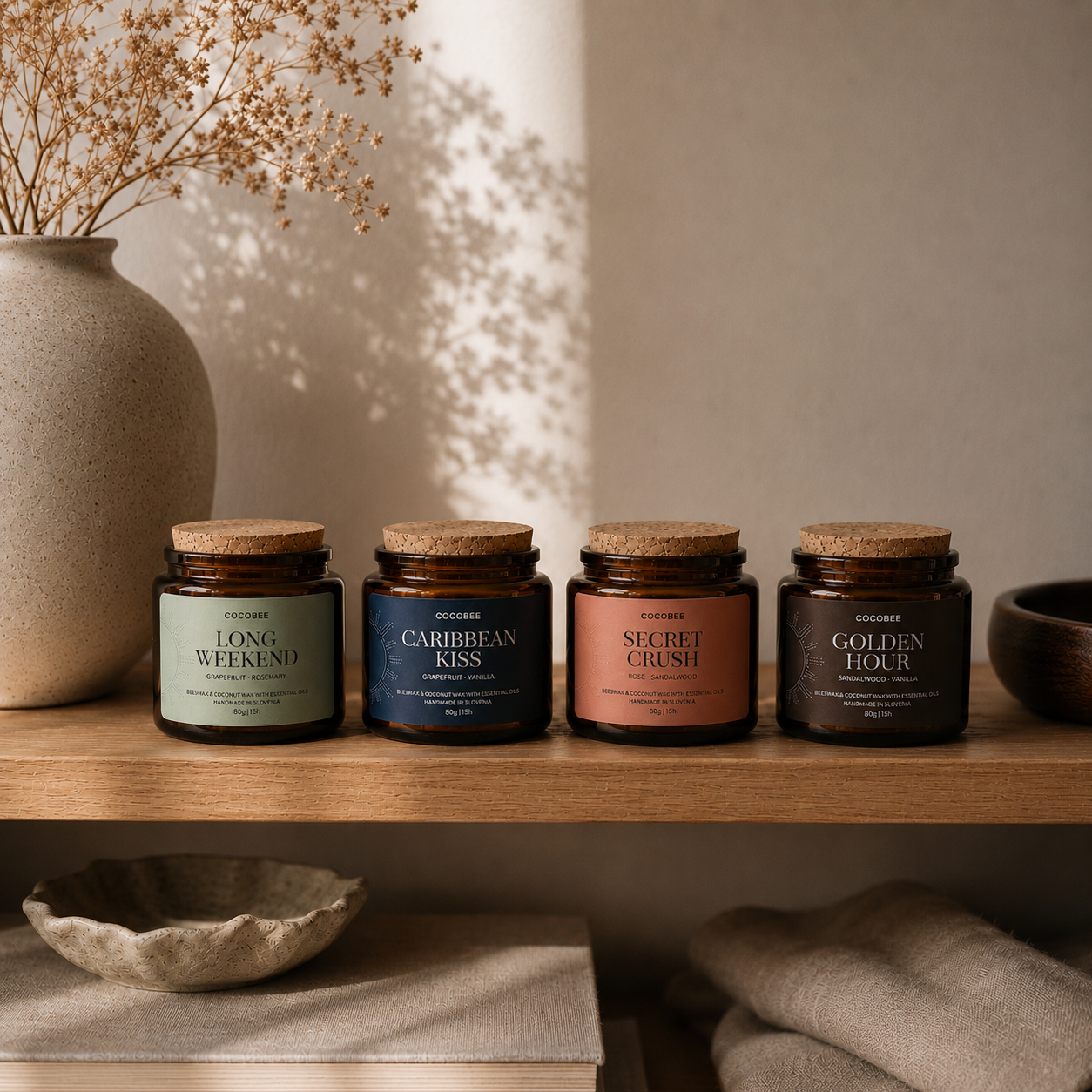Four candles with cork lids on a wooden shelf, with a vase and dried plants in the background.