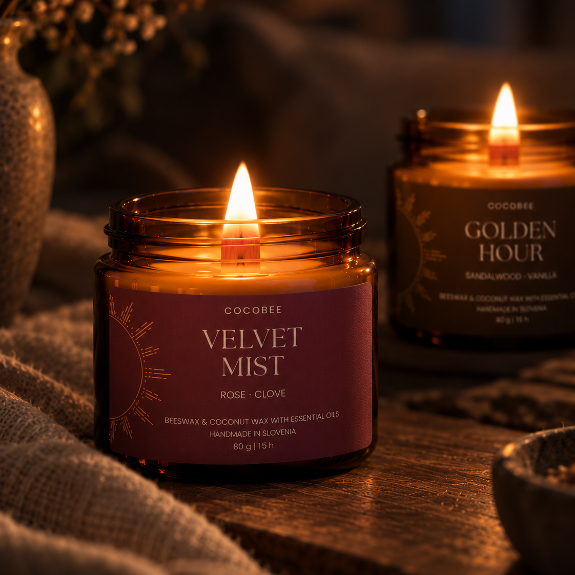 Two Cocobee candle jars on a rustic wooden surface with a linen cloth and dried flowers in the background. The foreground jar is labeled 'Velvet Mist' and is lit, while the background jar is labeled 'Golden Hour' and also lit.