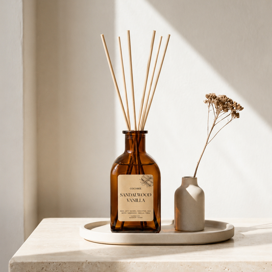 Amber glass reed diffuser bottle with wooden diffuser sticks on a round tray beside a small ceramic vase with dried flowers, set on a stone table in warm natural light.
