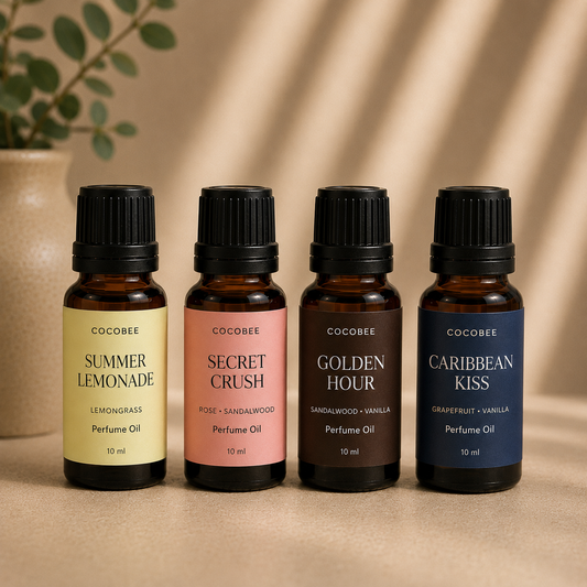 Four Cocobee perfume oil bottles lined up on a beige surface with a soft, warm background. The bottles are amber glass with black caps and feature labels in yellow, pink, dark brown, and navy blue, representing the Summer Lemonade, Secret Crush, Golden Hour, and Caribbean Kiss scents.