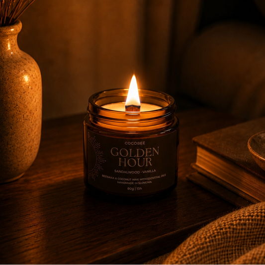 A lit candle in a dark amber glass jar sits on a wooden table in a cozy room setting, with a vase of dried stems on the left and a book on the right. The label reads 'GOLDEN HOUR'.