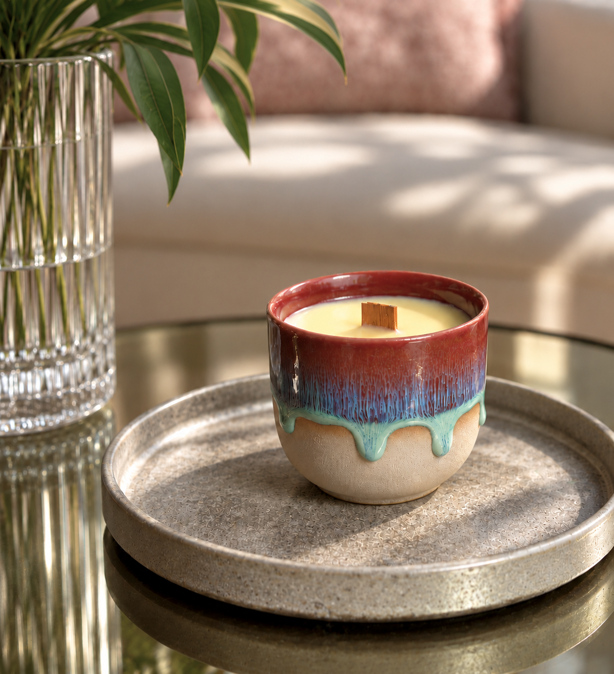 A ceramic candle in a small glazed cup sits on a round tray on a glass table, with a blurred living room background and a vase of green leaves at the side.