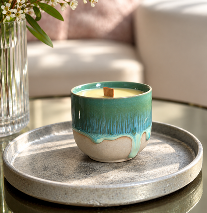 A ceramic candle in a small cup-shaped vessel with a teal-to-cream drip glaze and a wooden wick, displayed on a round gray tray in a softly lit interior setting.
