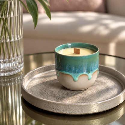 A ceramic candle in a small cup-shaped vessel with a turquoise and cream drip glaze, placed on a round tray on a glass table in a softly lit interior setting.