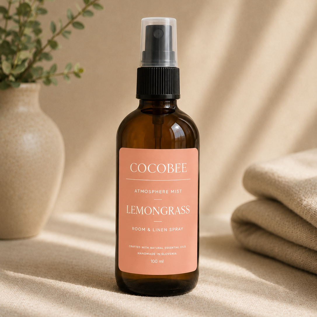 A bottle of Cocobee Lemongrass Room & Skin Spray in an amber glass spray bottle with a pink label, styled on a beige surface with soft shadows, a potted plant, and folded fabric.