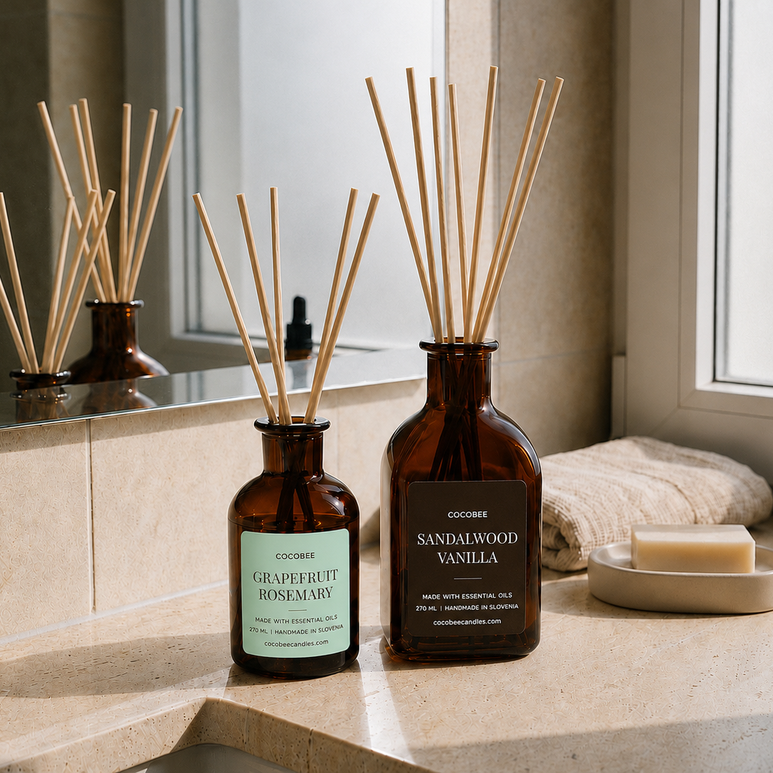 Two reed diffusers in amber glass bottles with wooden reeds, labeled Grapefruit Rosemary and Sandalwood Vanilla, styled on a beige stone countertop with a mirror reflection, a folded towel, and a soap dish in a bright bathroom setting.