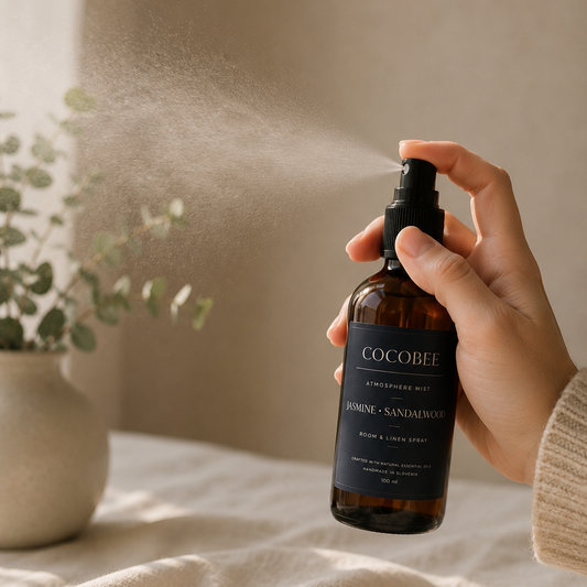 A hand holds a Cocobee home and garden room spray bottle, spraying a fine mist into the air. The amber glass bottle has a black spray top and a dark navy label with white text.