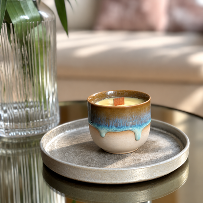 A small ceramic candle jar with a brown and blue glazed drip design sits on a round gray tray on a reflective table, with a glass vase and softly blurred sofa in the background.