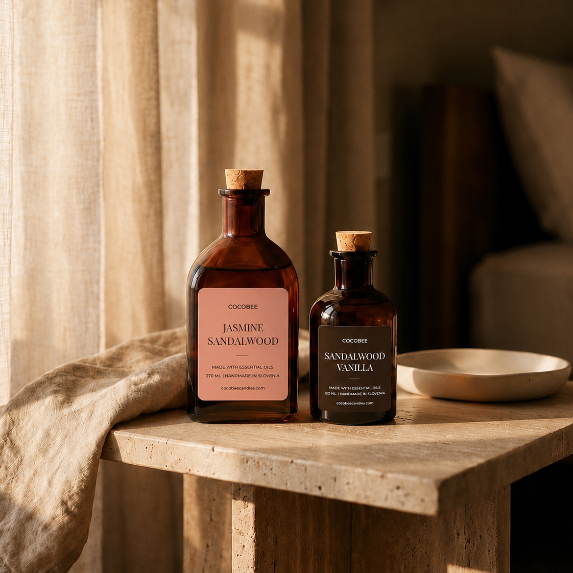 Two amber glass bottles of COCOBEE vanilla extract, one larger bottle labeled Jasmine Sandalwood and one smaller bottle labeled Sandalwood Vanilla, displayed on a rustic stone side table next to a linen throw and a ceramic dish in a warm, softly lit bedroom setting.