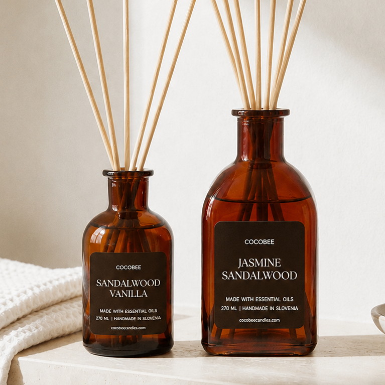 Two amber glass reed diffusers with natural wood reeds sit on a light stone ledge in a softly lit interior. The larger bottle is labeled Jasmine Sandalwood, and the smaller bottle is labeled Sandalwood Vanilla.