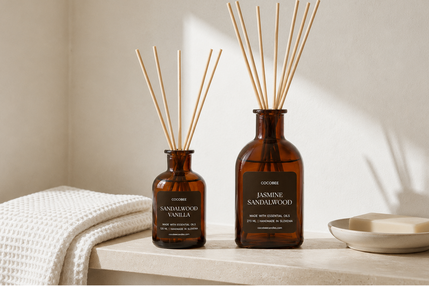 Two amber glass reed diffusers on a light stone surface, one labeled Sandalwood Vanilla and one labeled Jasmine Sandalwood, with natural reed sticks in each bottle and a folded white towel beside them.