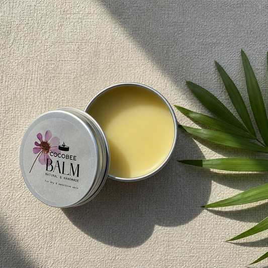 Cocobee balm tin with a lid open, showing a yellow balm inside, on a textured surface with a leaf.