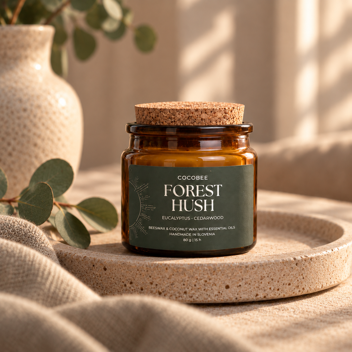 A glass jar candle with a cork lid and a dark green label reading 'COCOBEE FOREST HUSH' sits on a round stone tray, styled with linen fabric, eucalyptus leaves, and a ceramic vase in a warm neutral setting.