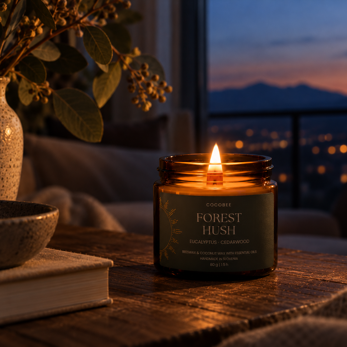 A lit candle in an amber glass jar sits on a wooden table in a cozy indoor setting at dusk, with a vase, books, and a city skyline visible in the background. The label reads 'COCOBBE FOREST HUSH'.