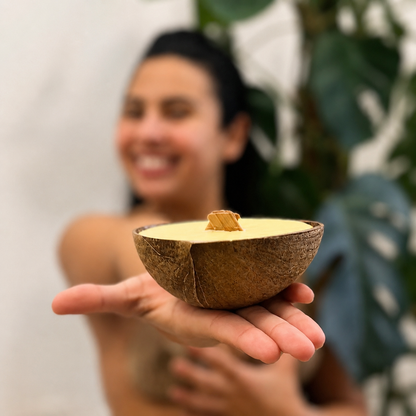 A person holding a coconut half bowl candle filled with beeswax and coconut wax and a wooden wick. The background is softly blurred with a white wall and green leaves.o