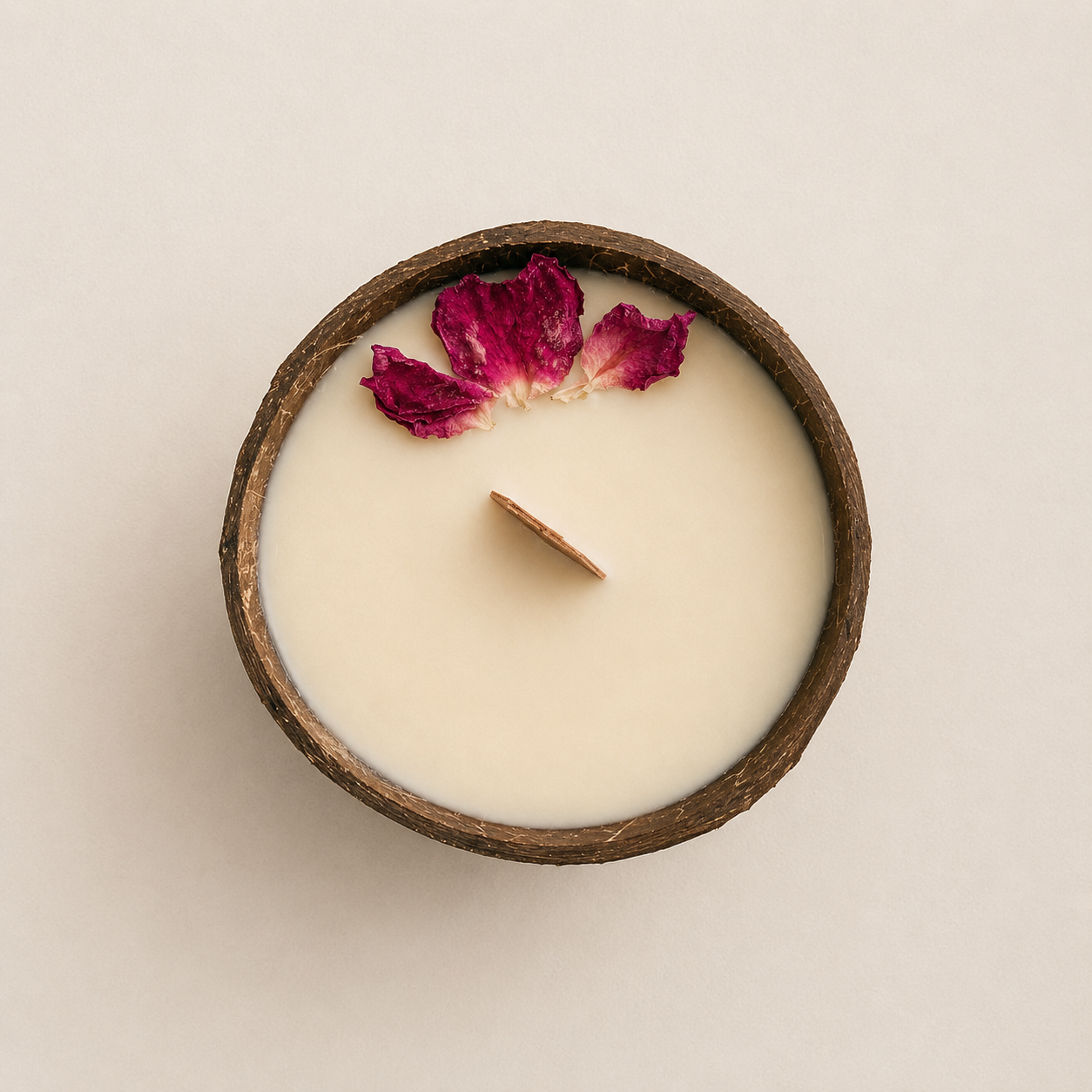 A top-down view of a round coconut shell candle with creamy beeswax and coconut wax, a wooden wick, and a dried magenta rose petal on the surface, set against a solid light beige background.