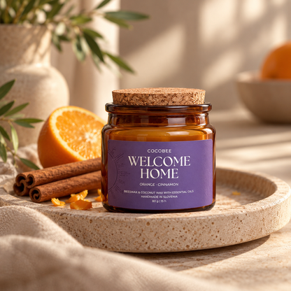 A Cocobee 'Welcome Home' scented candle in an amber glass jar with a cork lid, styled on a stone tray with cinnamon sticks and a halved orange.