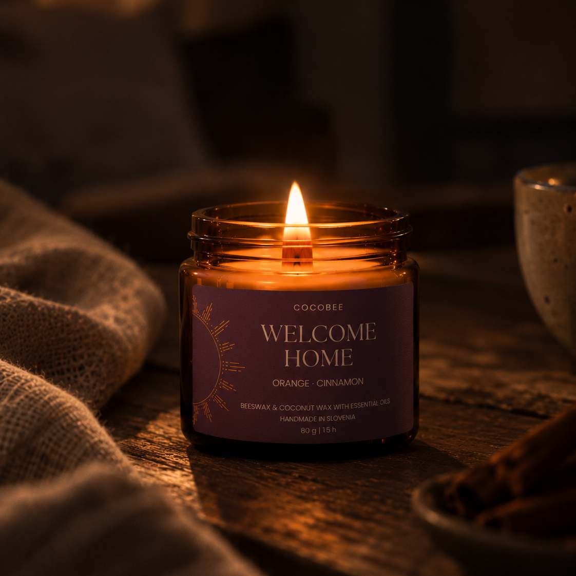 A lit candle in an amber glass jar with a purple label reading 'WELCOME HOME' and 'ORANGE - CINNAMON' by Cocobee, placed on a rustic wooden surface with warm ambient lighting and cozy props in the background.