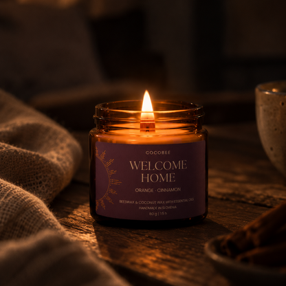A lit candle in an amber glass jar with a purple label reading 'WELCOME HOME' and 'ORANGE - CINNAMON' by Cocobee, placed on a rustic wooden surface with warm ambient lighting and cozy props in the background.